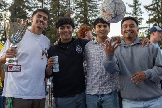 Four students standing at a table holding up a trophy and a ball in the air