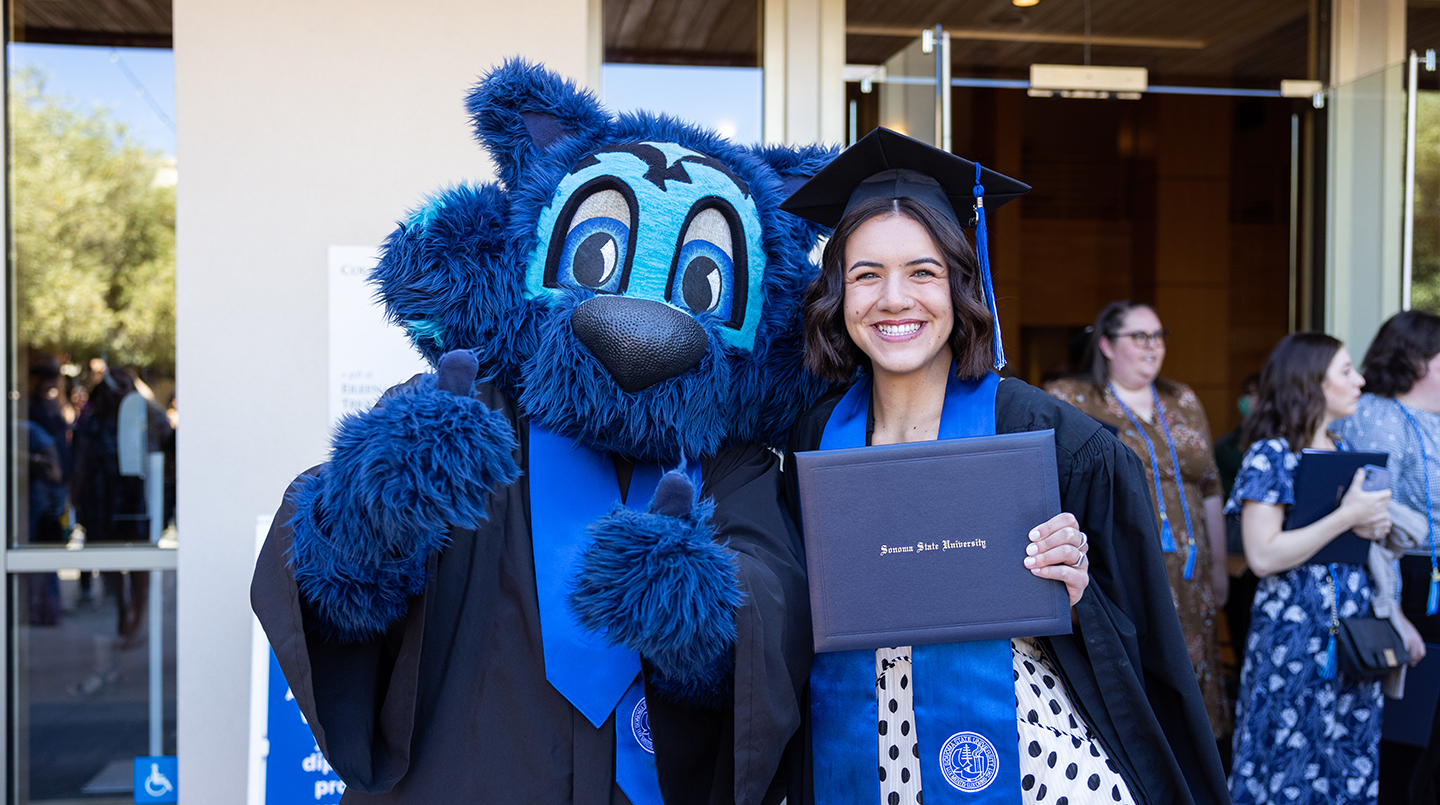 Graduate in cap and gown holds diploma,  next to Lobo mascot