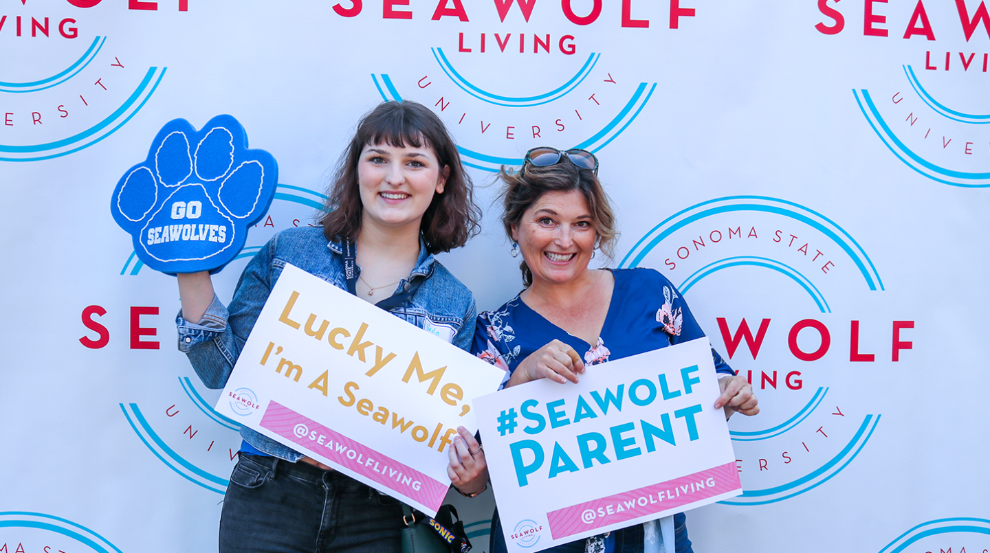 Two people holding signs that say "Lucky Me, I'm a Seawolf" and #Seawolf Parent"