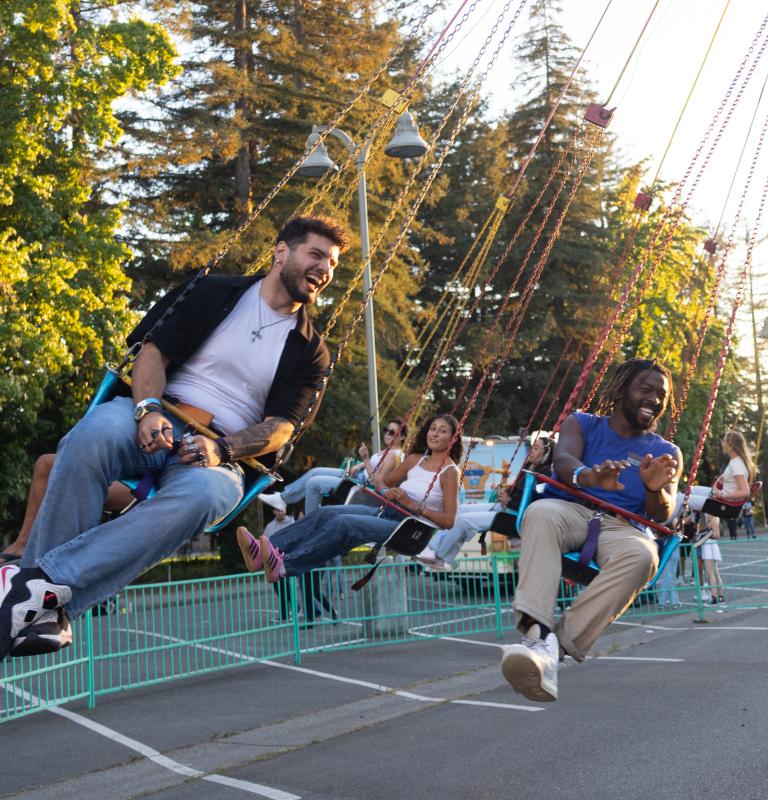 Two Sonoma State students laughing while riding a swing carousel at an outdoor fair during golden hour