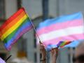 Hands holding up the rainbow LGBTQIA Pride flag as well as the pink, blue, and white Trans Pride flag