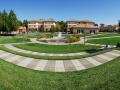 A wide-angle view of the outdoors area of on-campus housing including grass areas, trees, walking paths, and a fountain