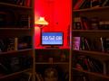 A darkly lit room with filled bookshelves, a red lamp, and a computer screen with the digits '00:52:08' displayed on it