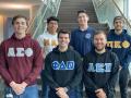 A group of six fraternity members standing in two rows and smiling while wearing different fraternity sweatshirts 