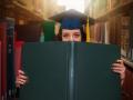 Person inside a library holding a large green diploma in front of their face while wearing a blue graduate cap with a yellow tassel 