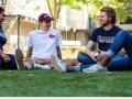 Four students smiling, talking, and sitting in the grass outdoors 