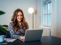 Someone smiling while sitting at an indoor table with a laptop in front of them and a mug, cellphone, and paperwork to their side