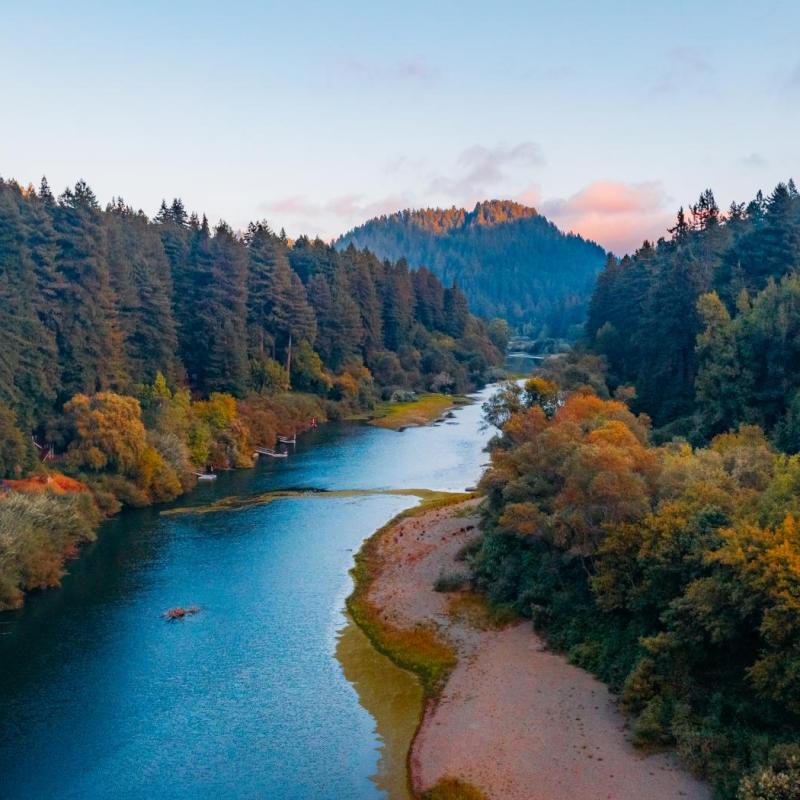 Russian river flows through a forested valley at sunset, with tree-covered hills and mountains in the background.