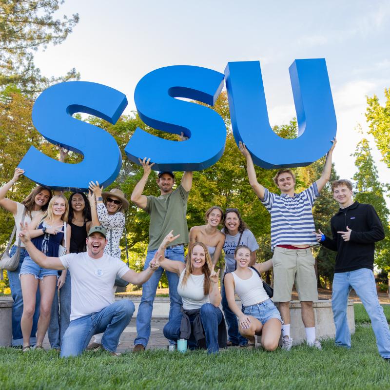 A group of smiling students pose on a grassy campus lawn holding large blue “SSU” letters above their heads, with trees and campus buildings in the background.