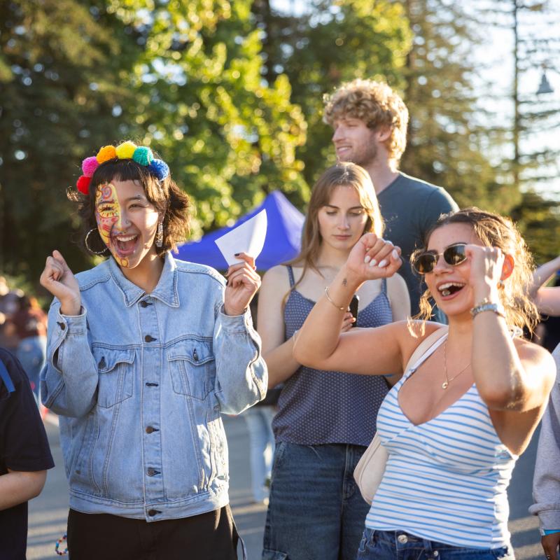 A group of students celebrate at an outdoor campus event, smiling and cheering, with one student wearing colorful face paint and a pom-pom headband in the center.
