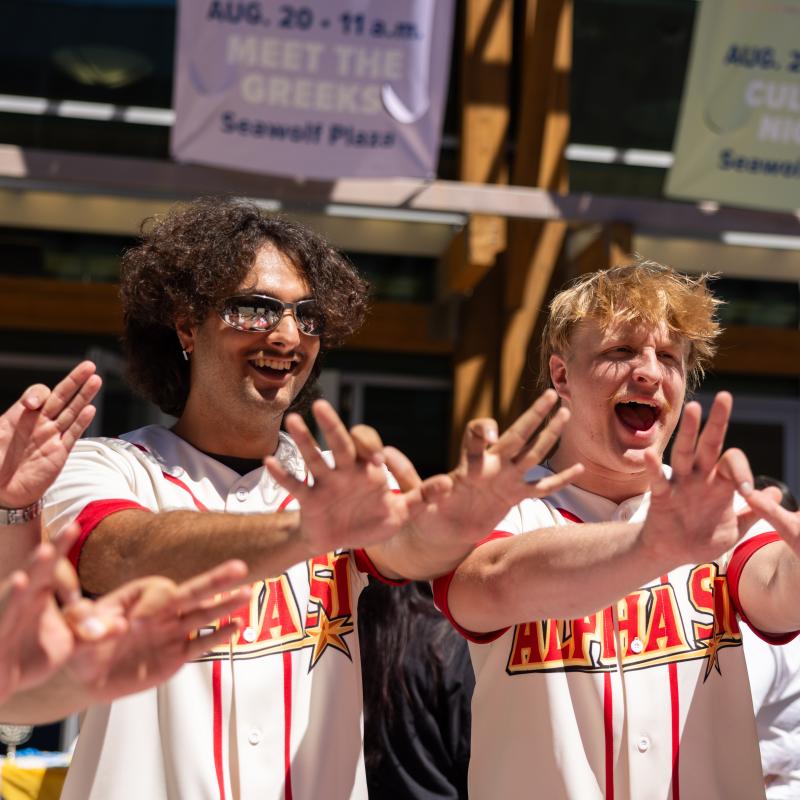 Three students wearing Alpha Sigma Phi jerseys stand outdoors on campus, smiling and extending their hands forward in a group gesture during a campus event.