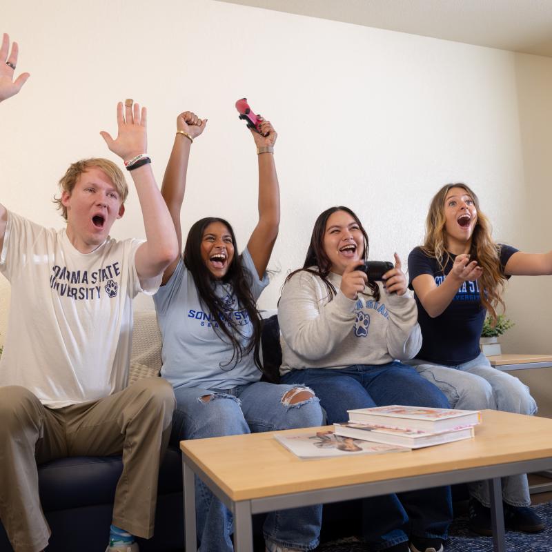 Four students wearing Sonoma State University shirts sit on a couch cheering and celebrating while playing a video game in a campus lounge.