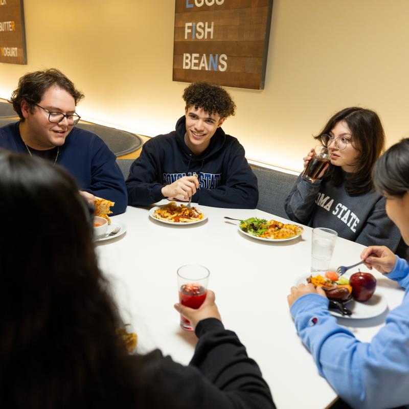 Five students wearing Sonoma State apparel sit around a table in a campus dining area, eating and talking together.