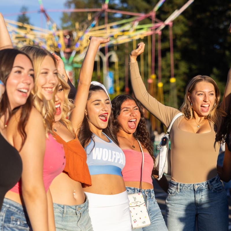 A group of students stand outdoors at a campus fair, smiling and cheering with their arms raised in front of a colorful swing ride.