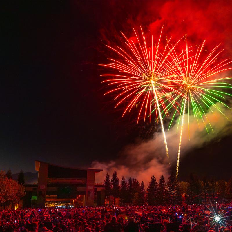 Red and green fireworks burst over a large nighttime crowd gathered on campus, with buildings and trees silhouetted beneath the illuminated sky.
